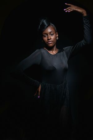 Elegant Black woman in black dress poses dramatically against dark background with raised arm.