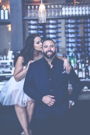 Woman in white dress kisses smiling bearded man in suit at upscale bar setting.