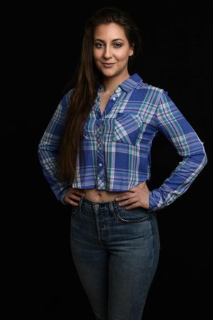 Young woman in blue plaid cropped shirt and jeans poses confidently against black background.