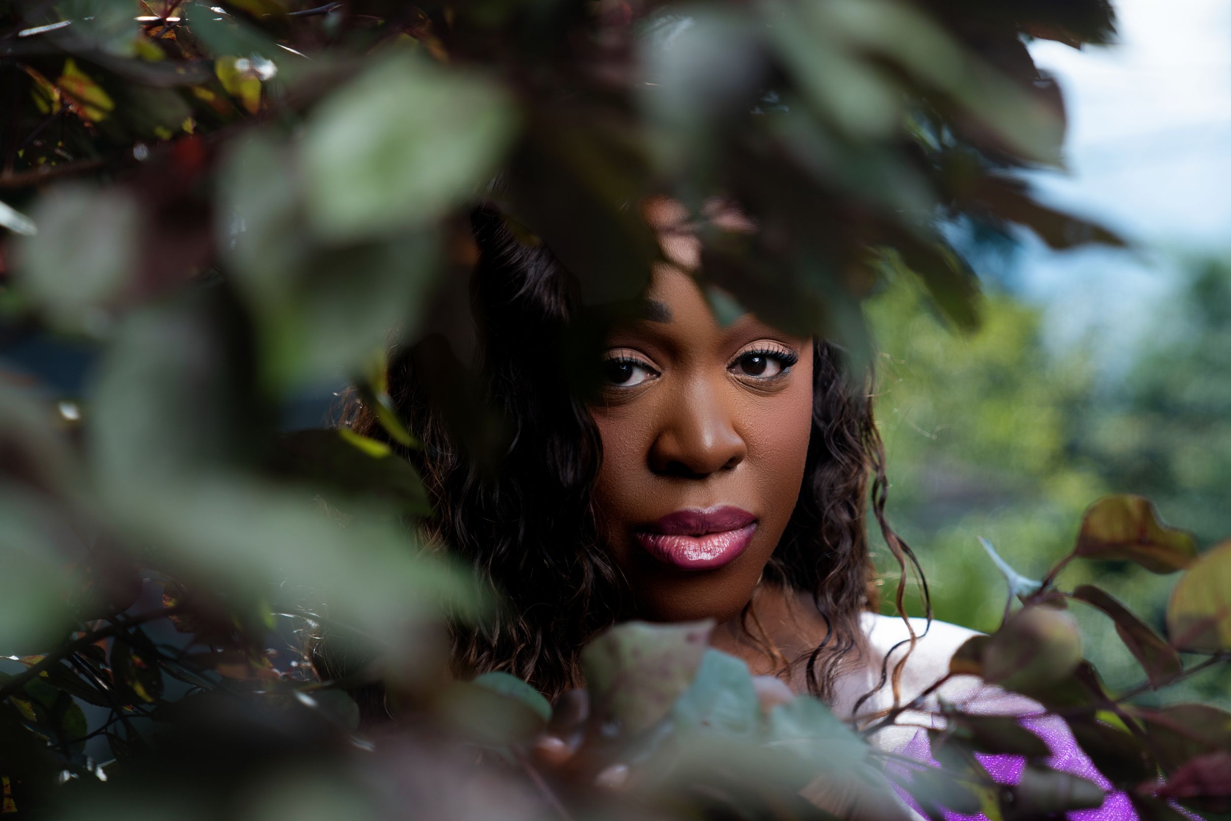 A Black woman peers through lush green and purple foliage, her expression thoughtful and serene.