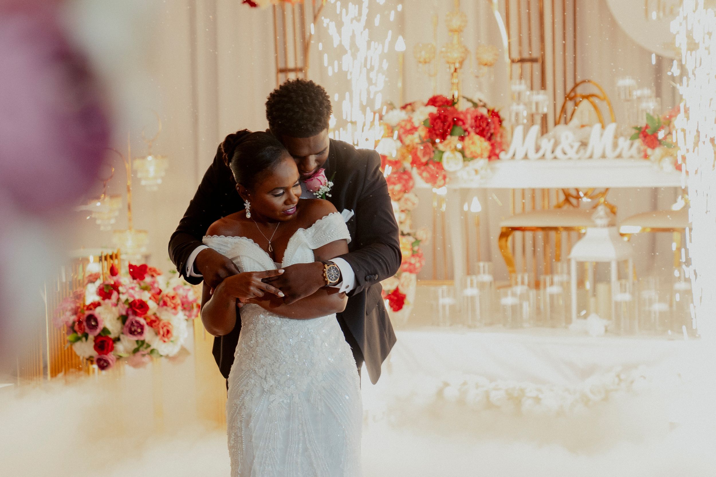Groom tenderly embracing bride in beaded gown during first dance at a lavishly decorated wedding reception.