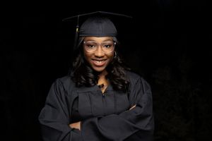 Proud graduate in black cap and gown smiles with arms crossed against dark background.
