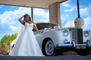 Bride in elegant white ball gown posing beside classic vintage white Rolls Royce luxury car.