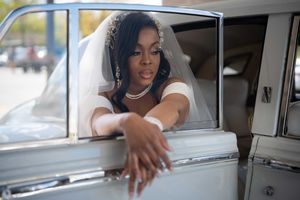 Beautiful Black bride in white wedding dress and veil leaning out of vintage classic car door.