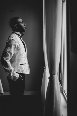 Black and white portrait of groom in ornate white tuxedo jacket gazing out window pensively.