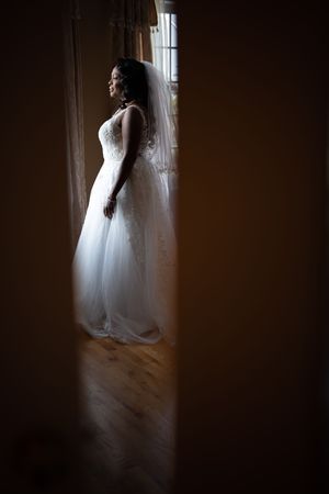 Bride in flowing white wedding gown and veil stands by sunlit window in dramatic silhouette.