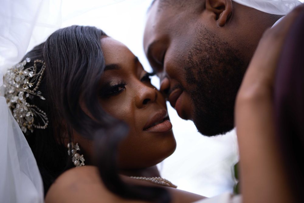Intimate close-up of newlywed couple nearly kissing under white veil with ornate bridal accessories.