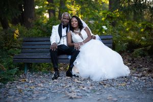 Newlywed couple in wedding attire sitting together on park bench surrounded by autumn foliage.