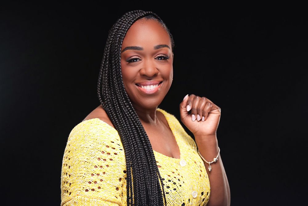 Smiling woman with long braids wearing yellow crochet top against dark background.