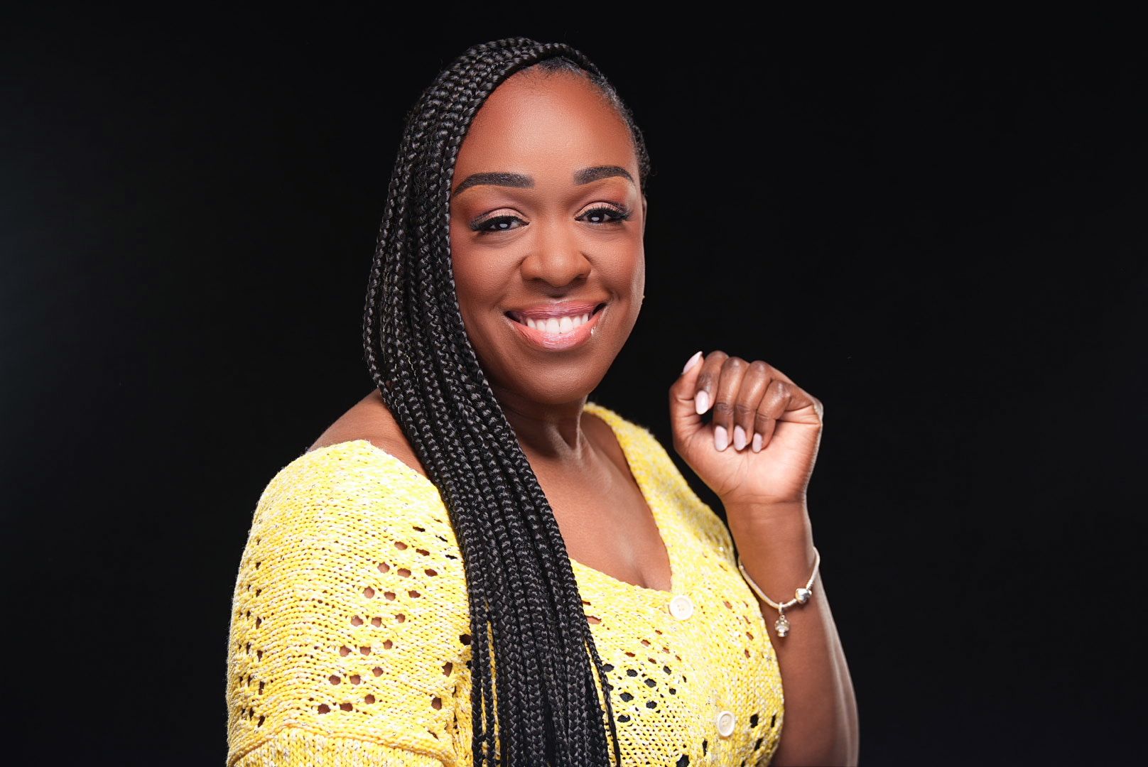 Smiling woman with long braids wearing yellow crochet top against dark background.