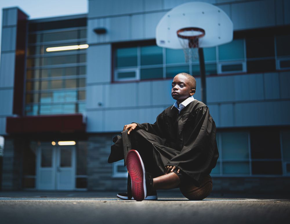 Graduate in black cap and gown sitting on outdoor basketball court in front of a school building.