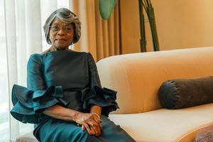Elegant older Black woman in teal ruffled dress seated on cream sofa in stylish interior setting.