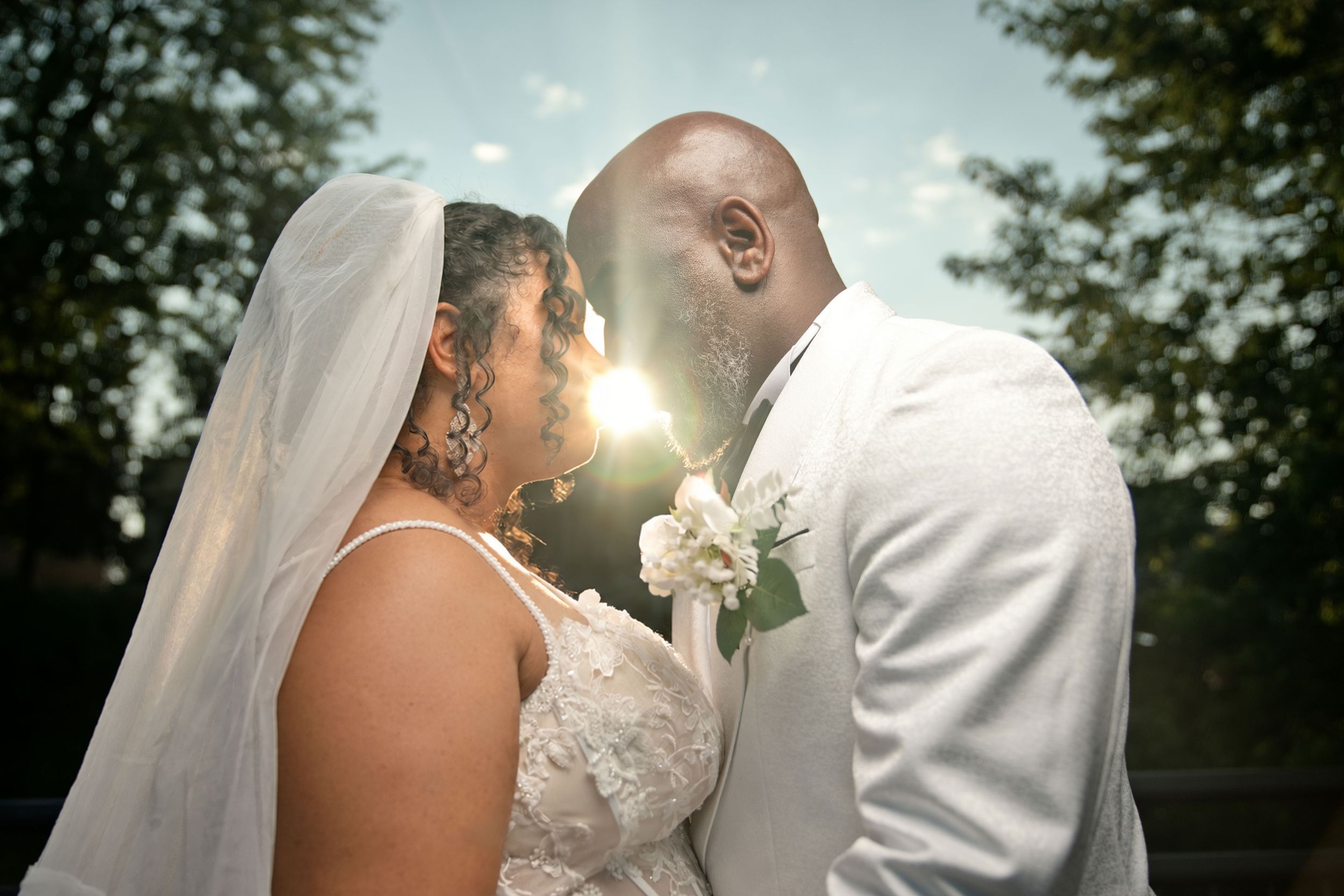 A bride and groom share an intimate moment, touching foreheads with sunlight glowing between them outdoors.