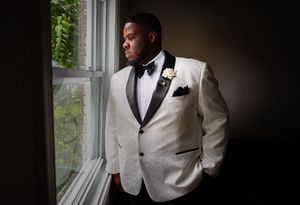 Black groom in white tuxedo jacket gazes out window in soft light.