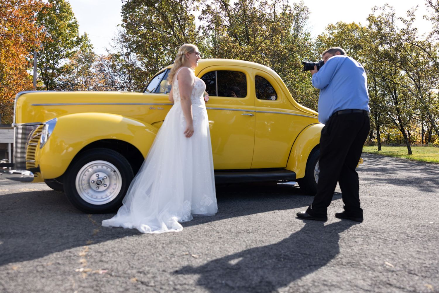 A yellow classic car serves as a backdrop for a wedding photoshoot with a bride in a flowing white dress.