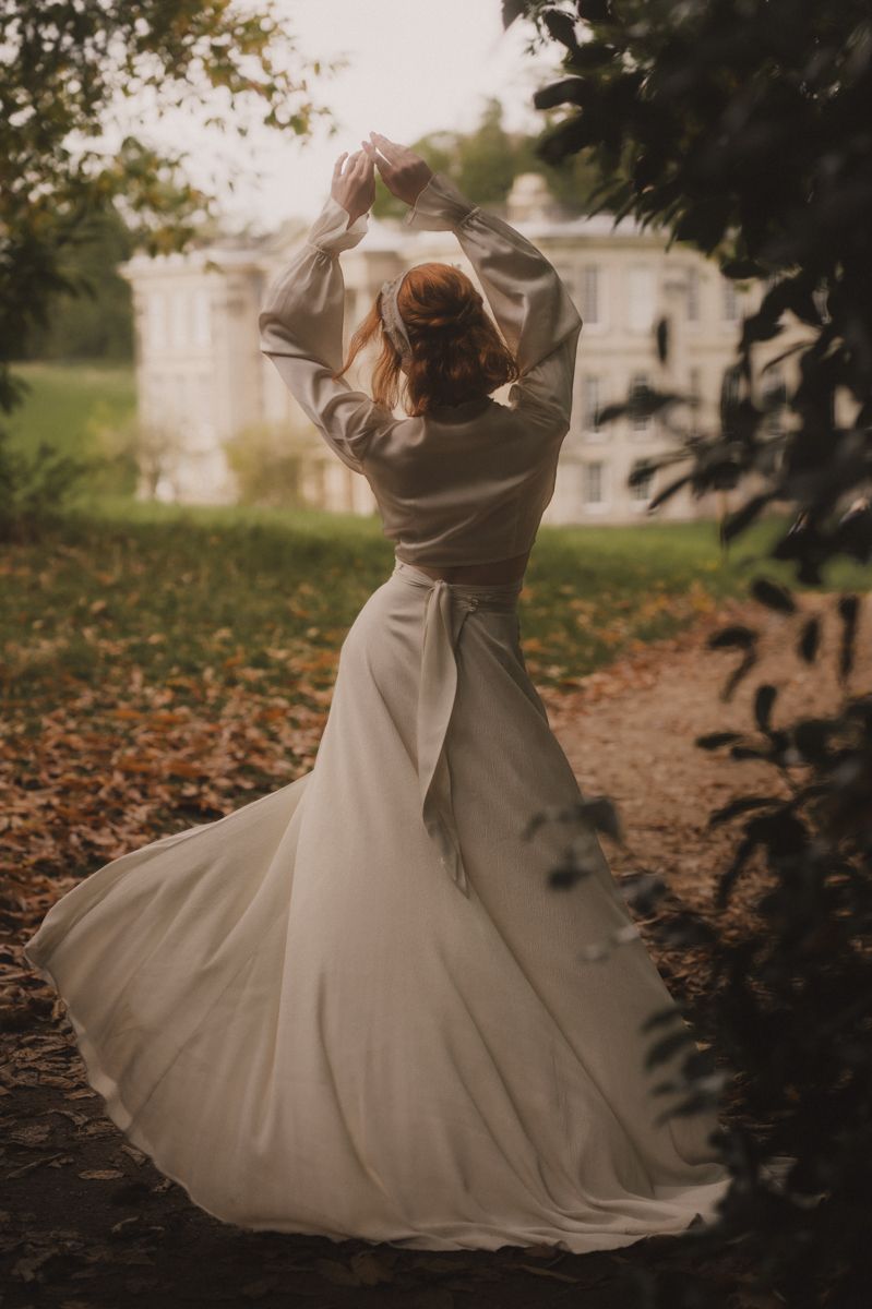 A person in a flowing white wedding dress twirls gracefully on an autumn path near a grand building at dusk.
