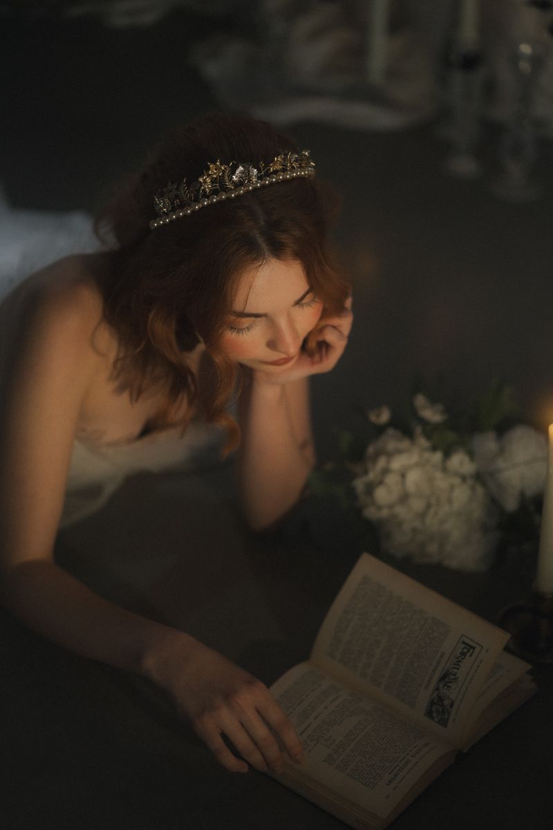 A person lies down while reading a book in dim lighting with white flowers visible in the background.