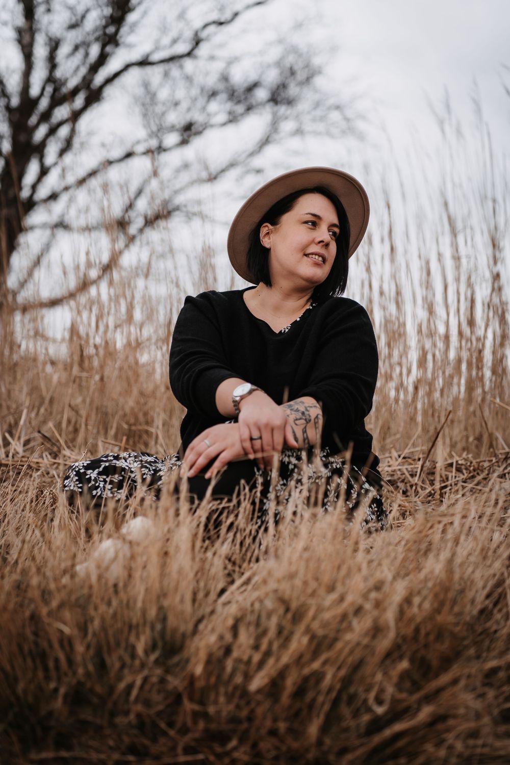 A candid portrait shot of someone wearing a brown hat sitting among dried grass in warm natural lighting.