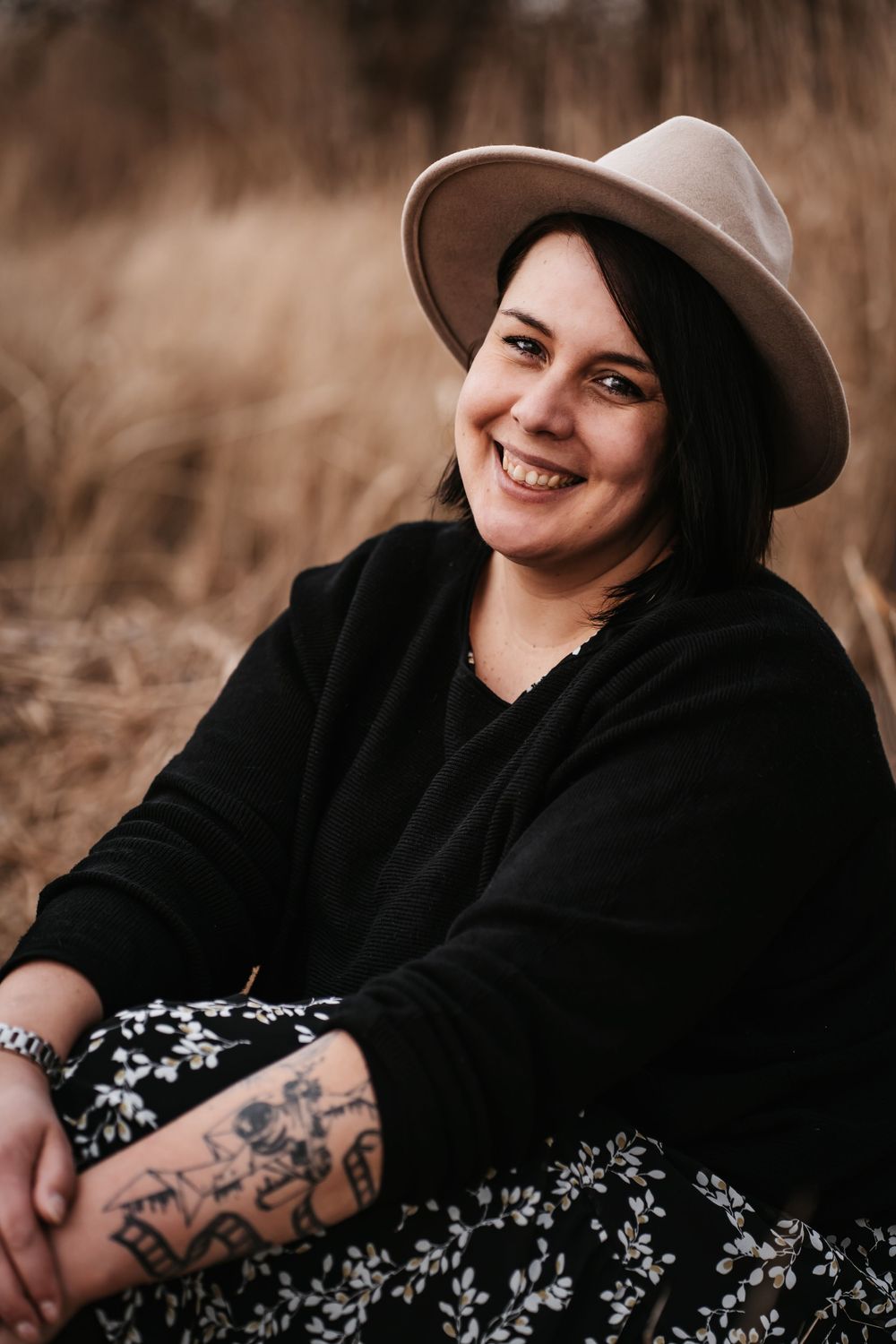 Person wearing a beige hat and black sweater sits outdoors in a floral dress with visible tattoos.