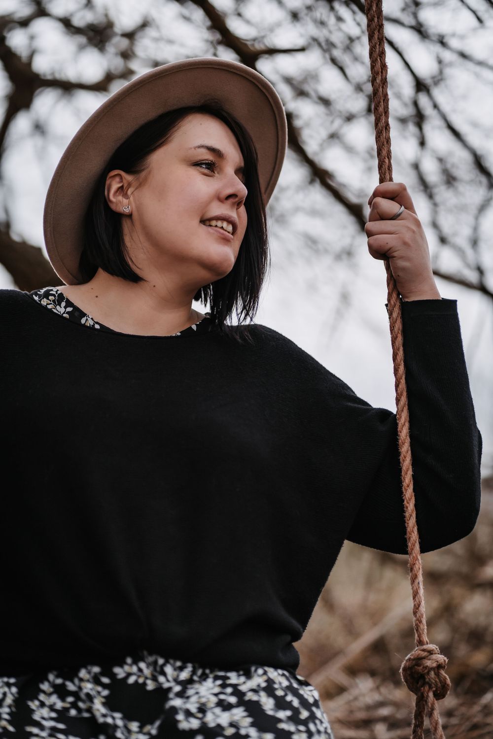 Person in a black sweater and brown hat holding onto a rope swing in a moody outdoor setting.