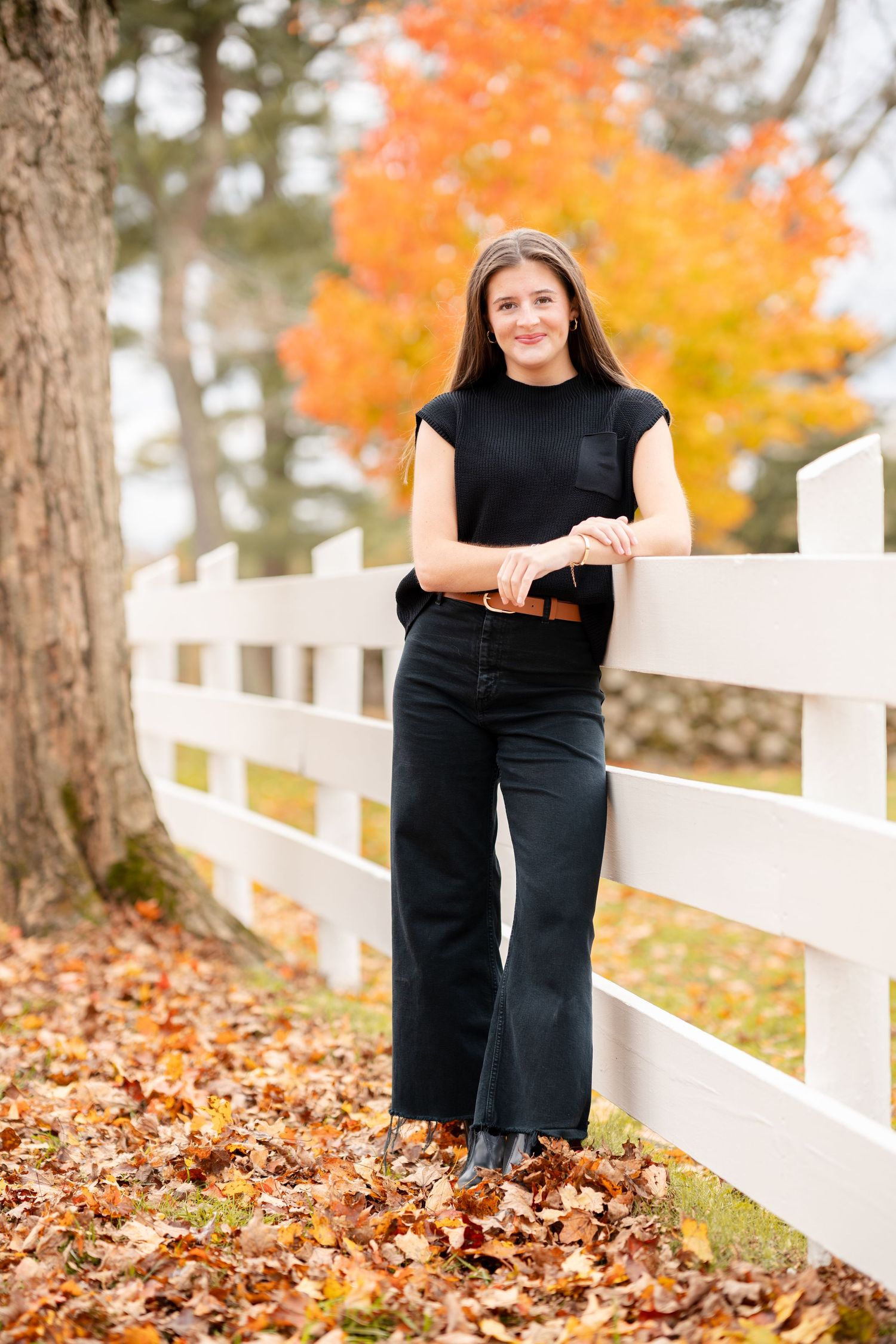 Person in black outfit poses near white fence with vibrant orange autumn foliage in background during fall photoshoot.