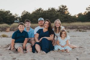 Family sitting together on sandy beach at sunset with pine trees in background.
