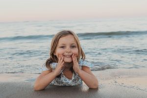 Child lying on beach sand with playful expression as waves roll in behind.