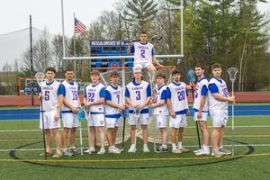 High school lacrosse team poses together on a blue turf field in white and blue uniforms holding sticks.