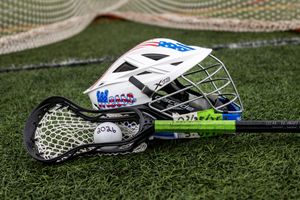 White lacrosse helmet with face guard resting on green turf beside lacrosse sticks and a white ball near a goal net.