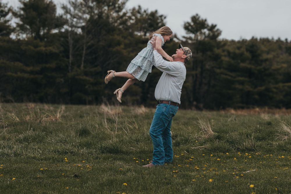 A father joyfully lifts his young daughter high in the air in a green field with trees in the background.