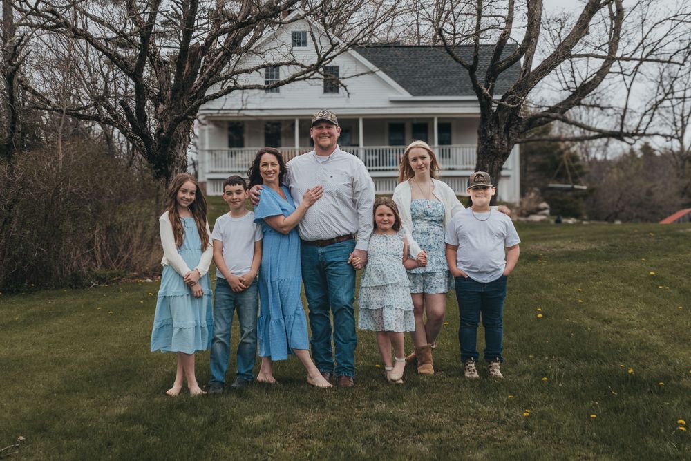 Family of seven posing outdoors in front of a white farmhouse, dressed in coordinating blue and white outfits.