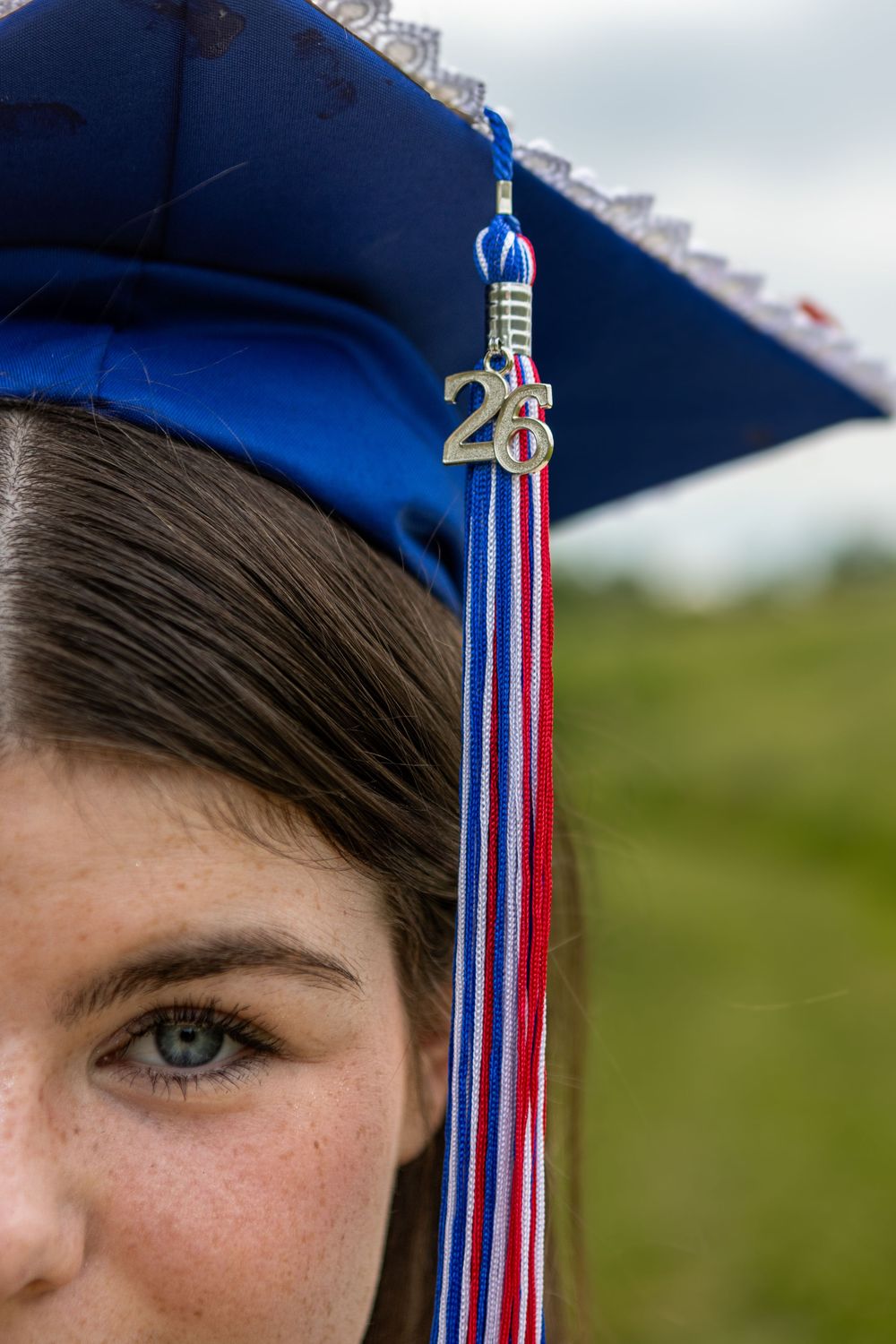 Close-up of a young woman wearing a blue graduation cap with a red, white, and blue tassel and gold 26 charm.