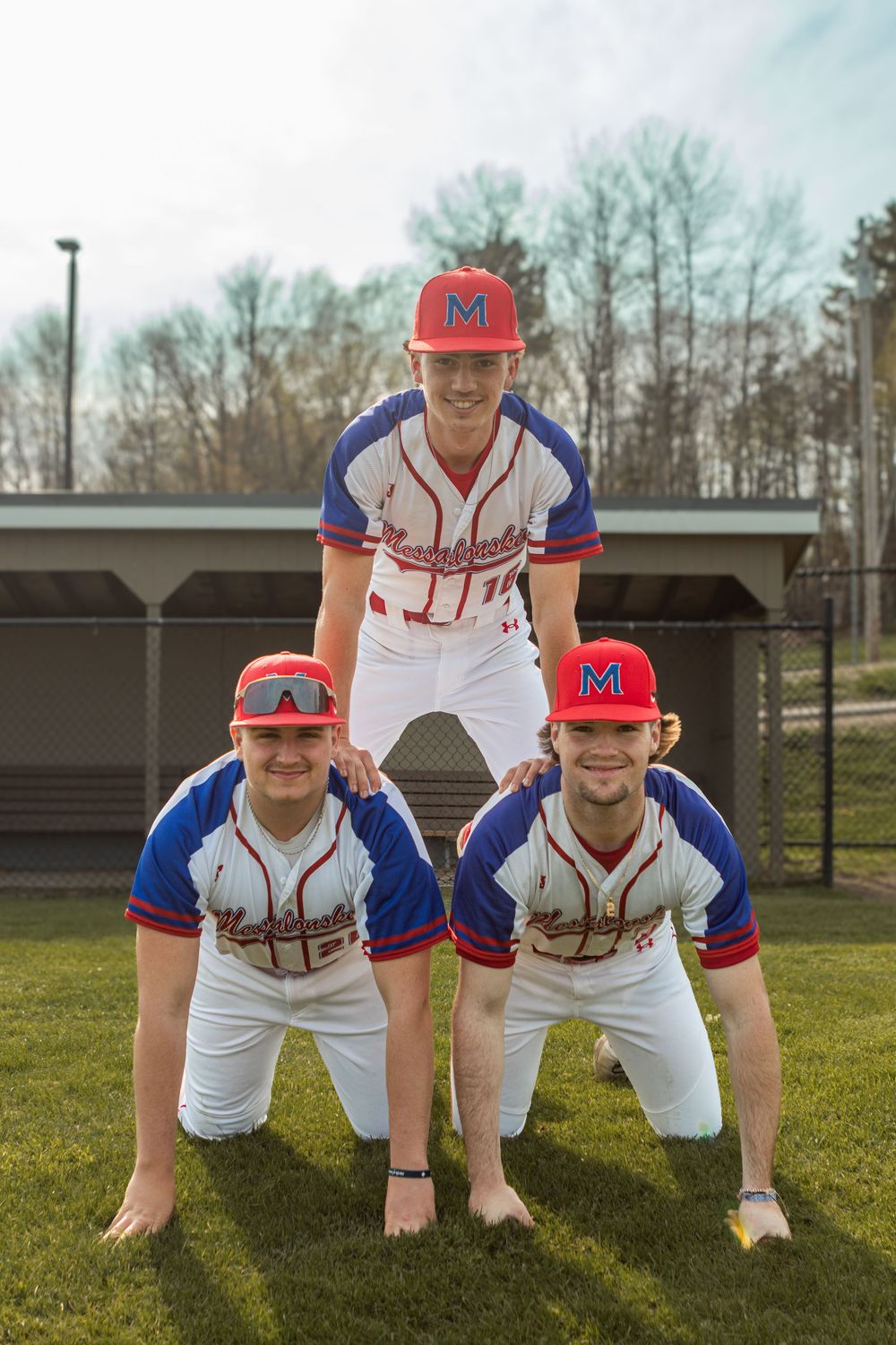 Three baseball players in red, white, and blue uniforms pose in a human pyramid on a green field.