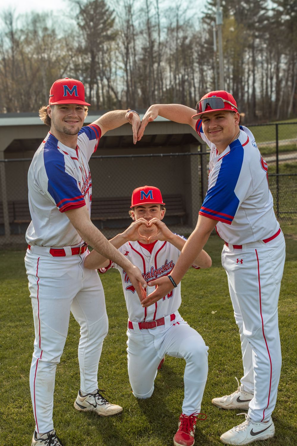 Three baseball players in red, white, and blue uniforms form a heart shape with their hands during a team photo outdoors.