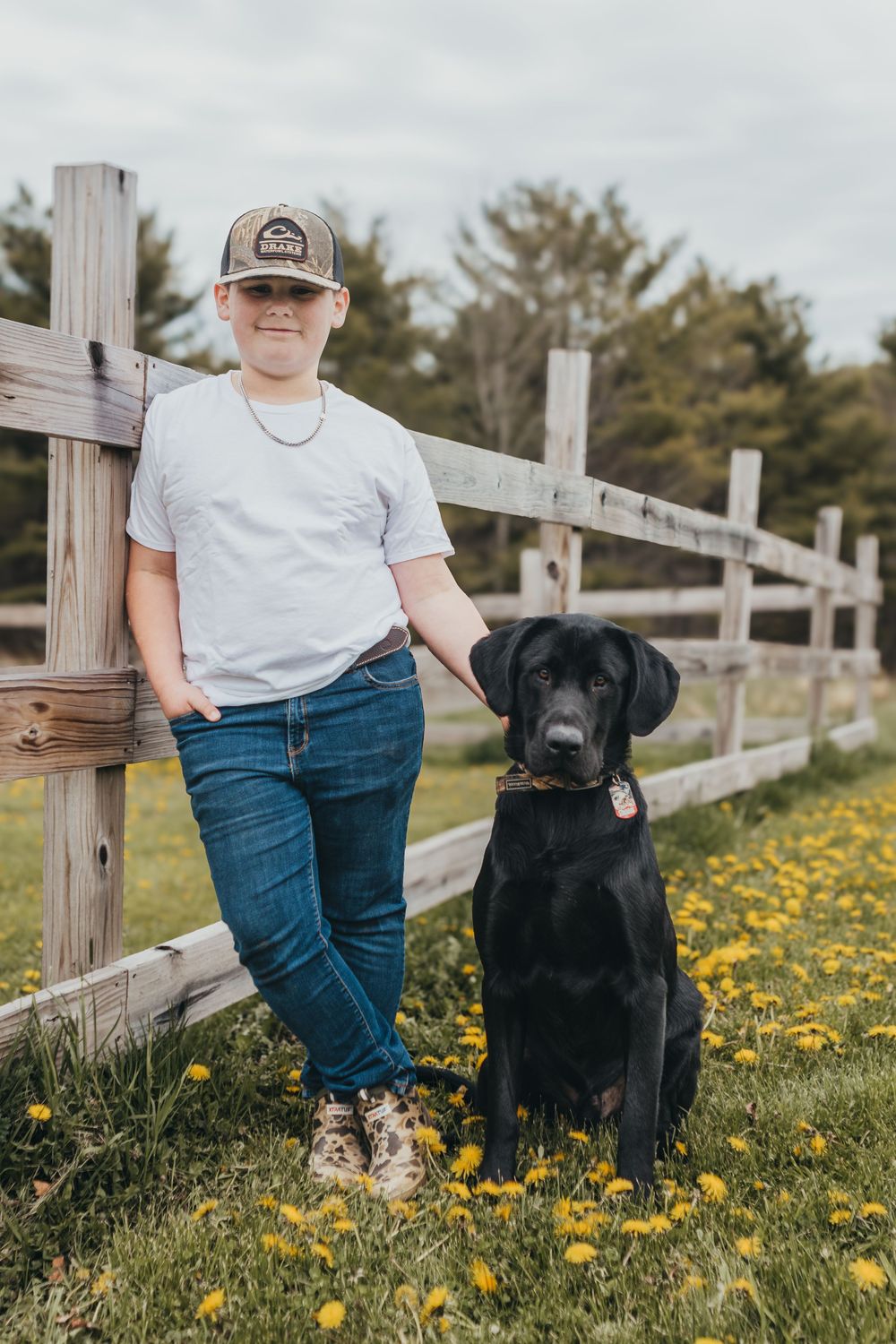 A young person in a cap and jeans poses with a black Labrador by a wooden fence in a field of dandelions.