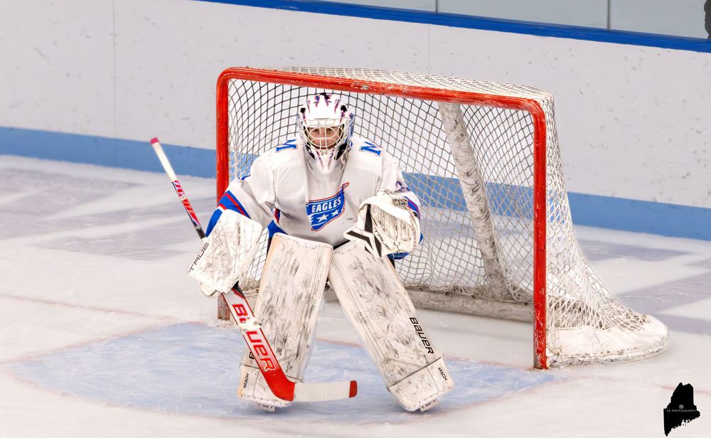 Ice hockey goalie in white and blue gear stands ready in front of a red net on an indoor rink.