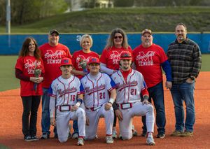 High school baseball seniors pose with family on field during senior night ceremony, wearing red and white uniforms.