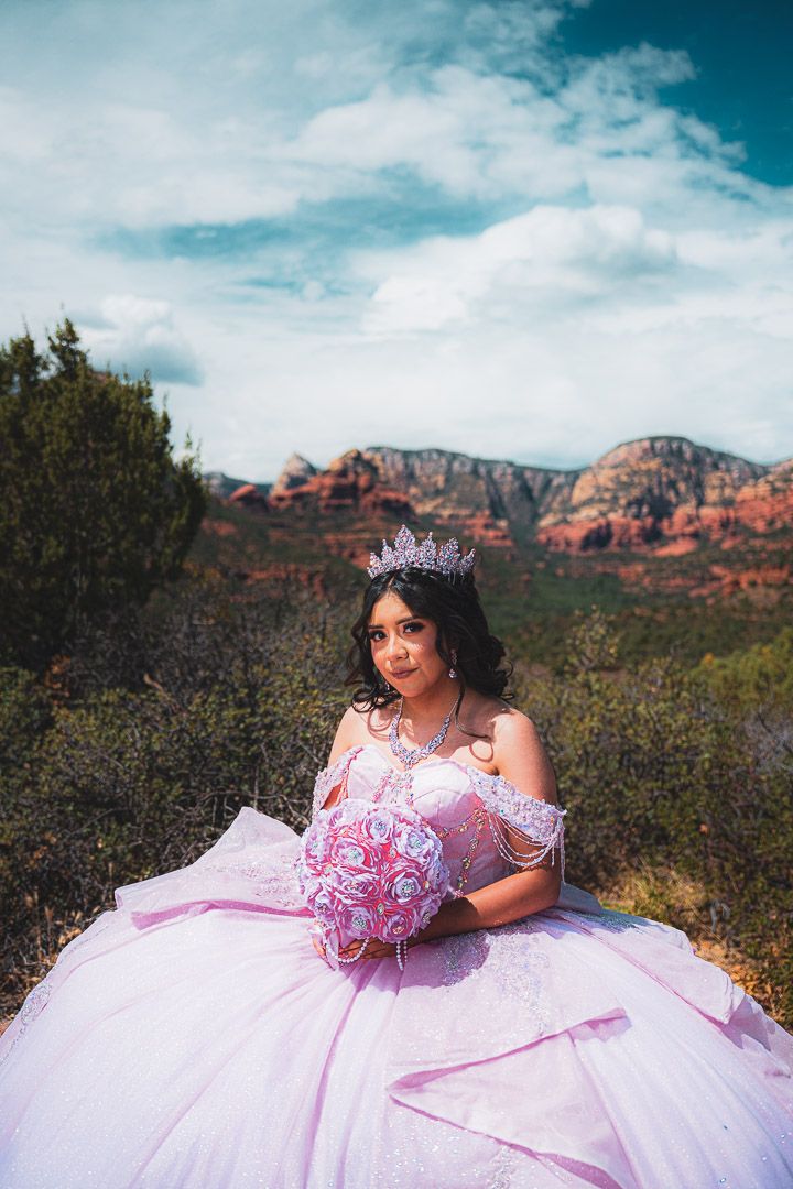 A person in a pink quinceañera dress and tiara poses against the backdrop of red rock mountains in Sedona, Arizona.