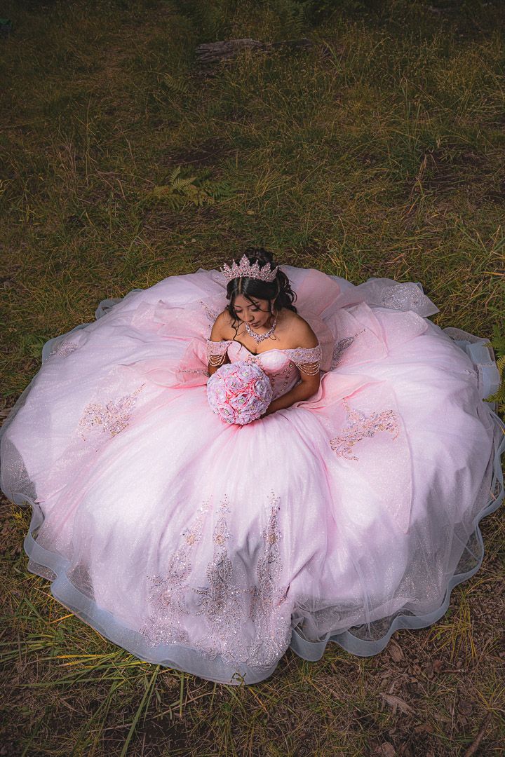 A pink quinceañera dress with delicate embroidery and tulle layers spreads out in a circular pattern on grass.