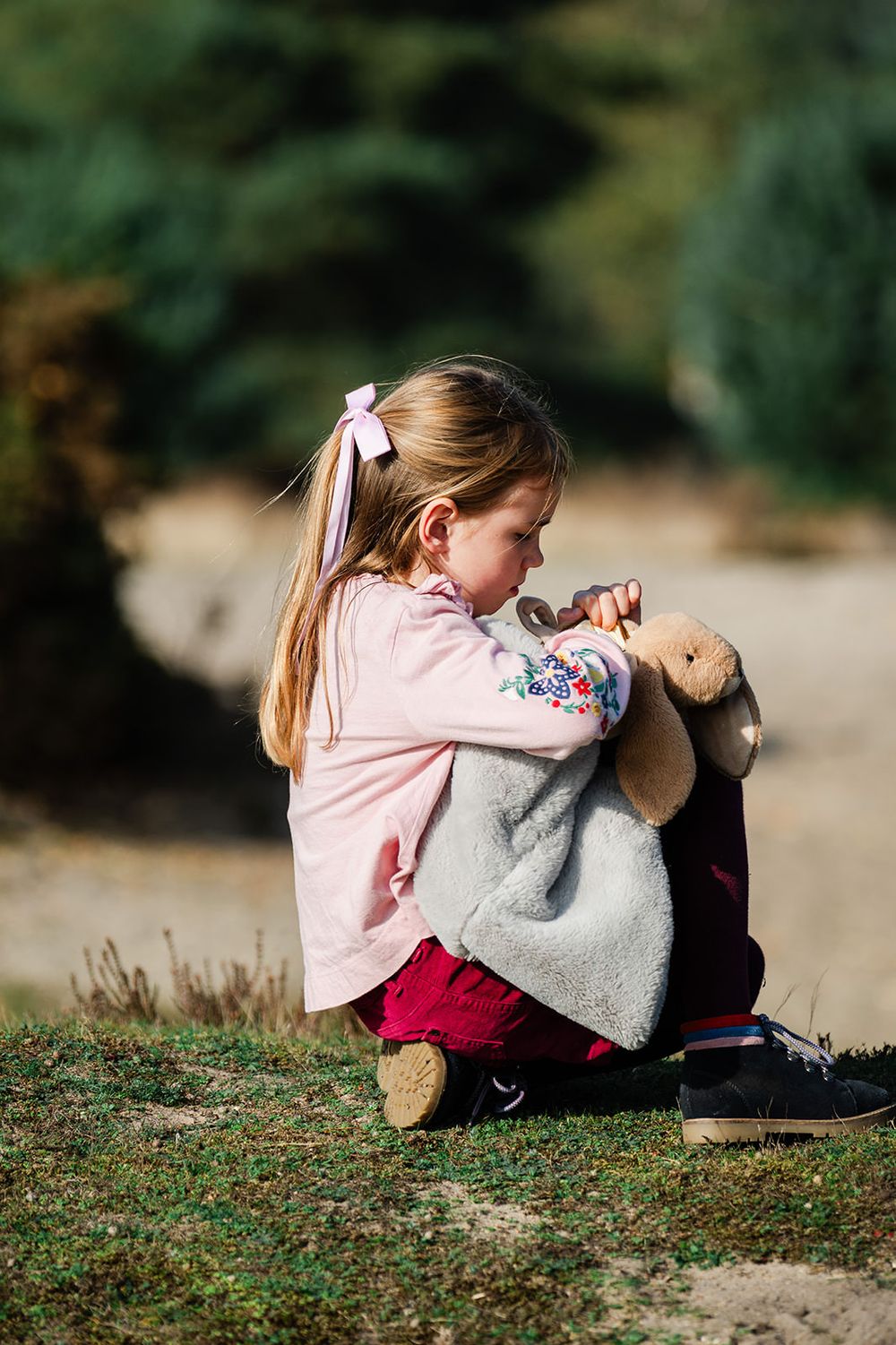 A child in a pink sweater sits outdoors holding a plush bunny toy on a grassy hill.