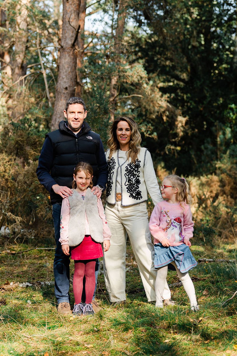 A family of four stands together outdoors in a wooded area on a sunny autumn day.