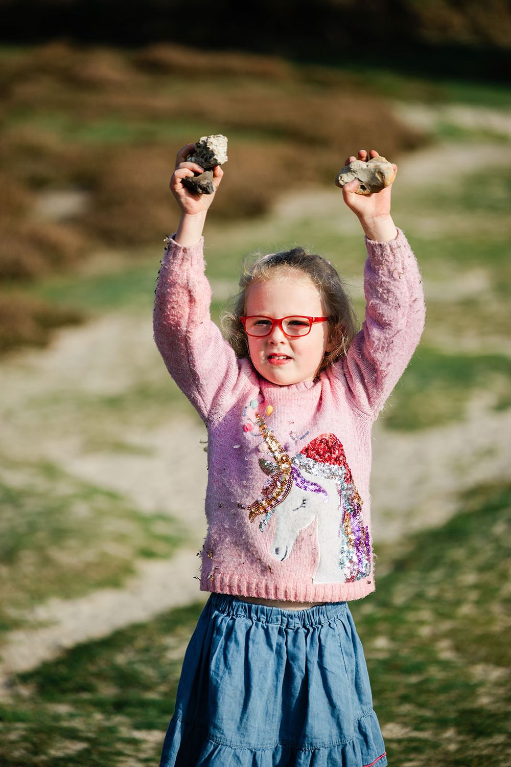 A person in a pink sweater and blue skirt raises their arms joyfully against a green outdoor background.