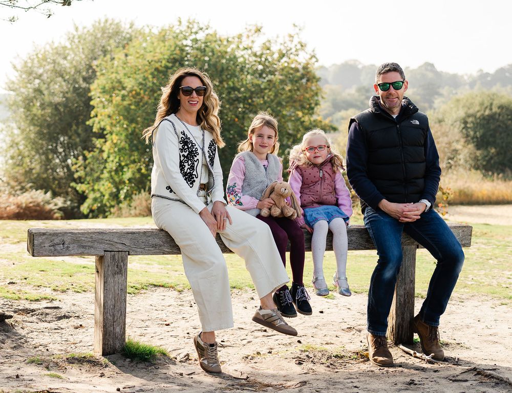 A family sits together on a rustic wooden bench outdoors on a sunny autumn day with trees in the background.