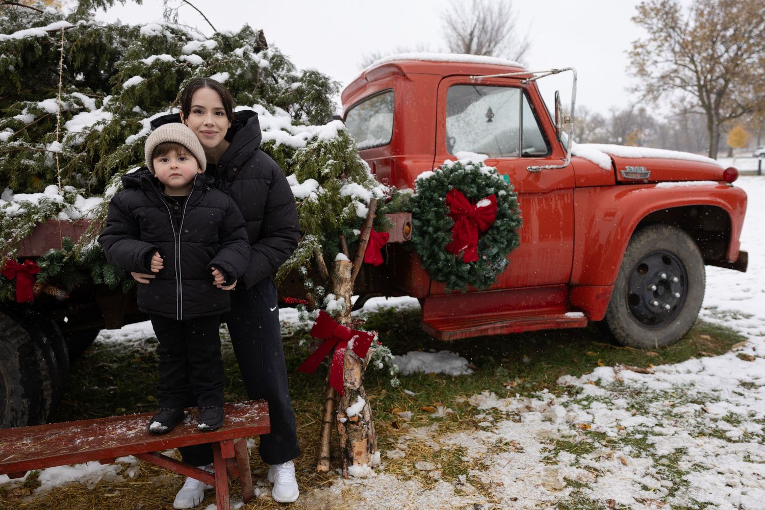 A woman and young child pose by a vintage red truck decorated with Christmas wreaths in snow.
