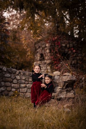 Child in burgundy velvet dress sits by rustic stone wall in autumn setting with golden light.