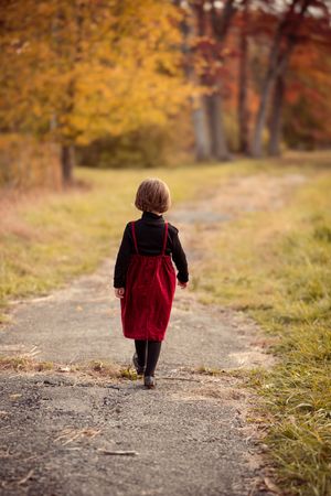 A small child in a red velvet dress walks alone on an autumn path surrounded by golden foliage.