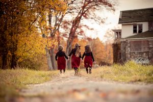 People in burgundy dresses walk along a scenic autumn path with golden foliage and rustic buildings in the background.
