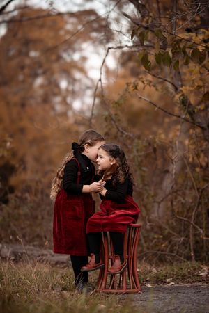 Two children in red velvet outfits share a tender moment in a fall forest setting with brown foliage.