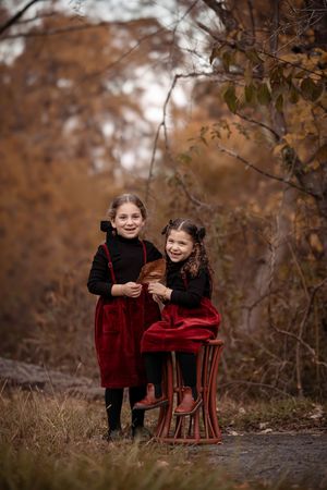 Two children in red velvet outfits share sweet moments on an antique chair outdoors during autumn.