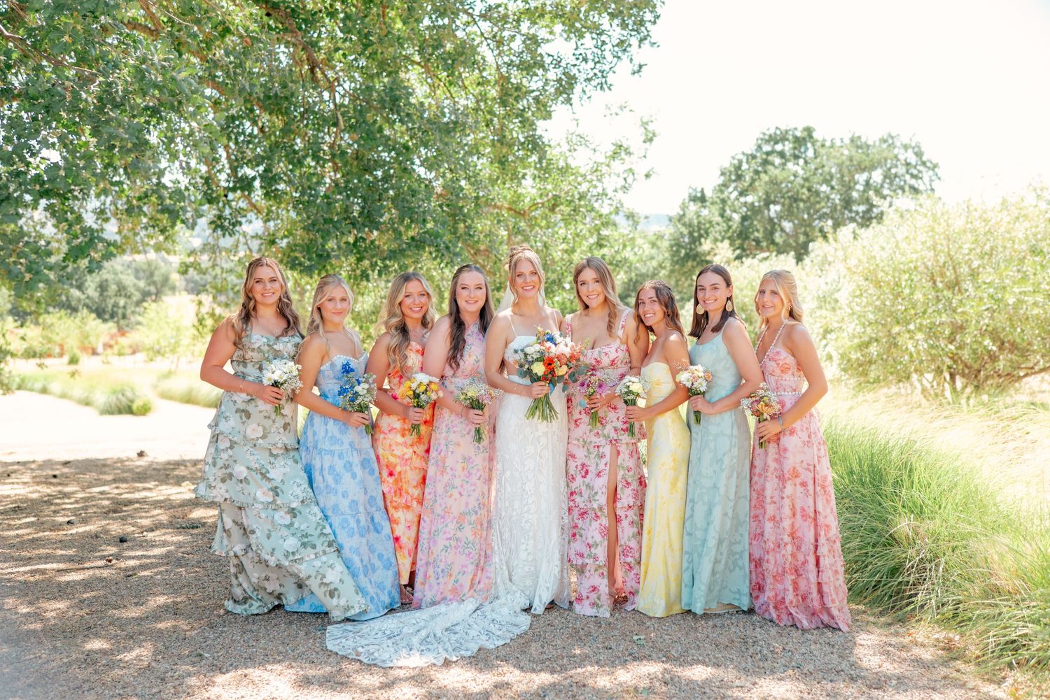 Bridesmaids in colorful floral dresses posed together outdoors under trees.